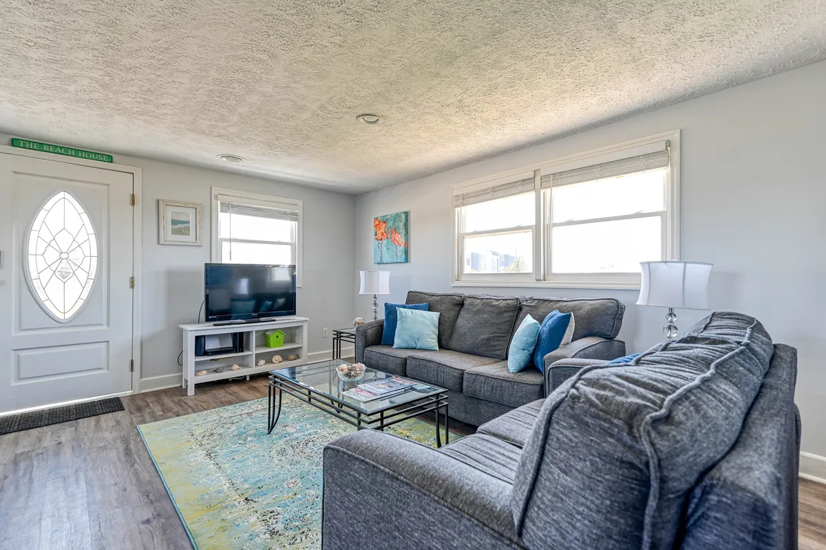 This image showcases the cottage’s main living area featuring a comfortable sofa, a coffee table, TV, and artwork near the front door. The inviting setup suggests a casual and relaxed atmosphere where guests can unwind after a day of beach or local adventures.