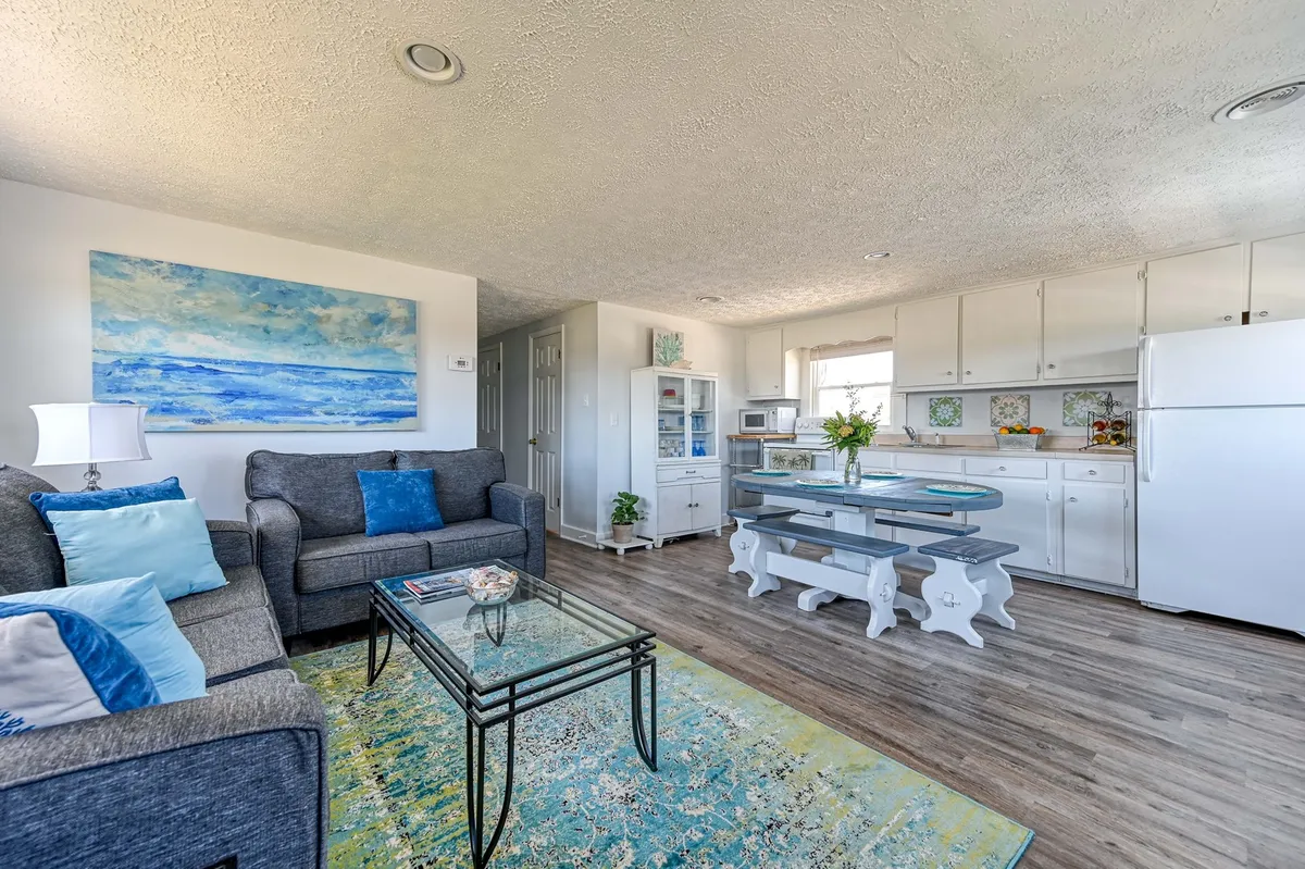 A bright, open-plan living and dining area with a comfortable sofa, stylish glass coffee table, visible kitchen space, eye-catching artwork on the walls, and a classic dining table by large windows that let in abundant natural light. The windows frame distant water views, reinforcing the coastal ambiance. The space feels vintage yet welcoming, perfect for gatherings or quiet relaxation.