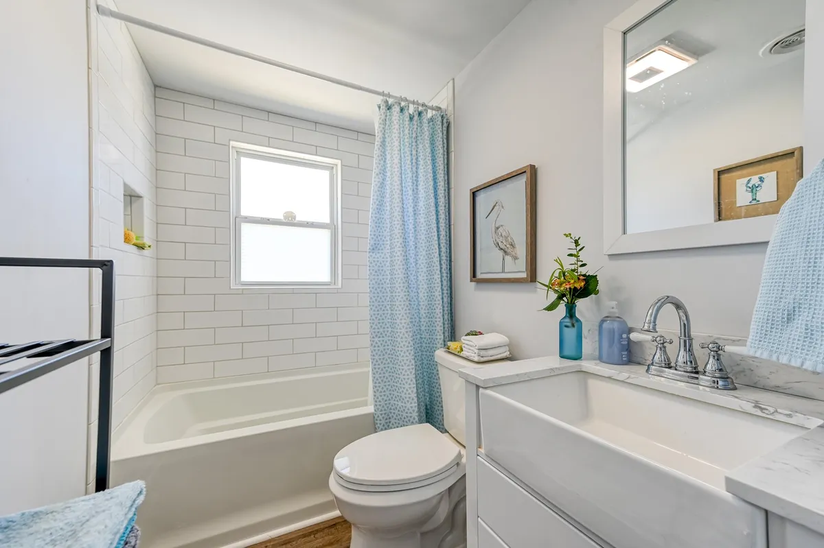 A bright, welcoming bathroom with a classic bathtub and farmhouse sink, framed artwork, a flower arrangement, and a sunlit window. The scene exudes calm and vintage elegance, ideal for daily routines or relaxing soaks.