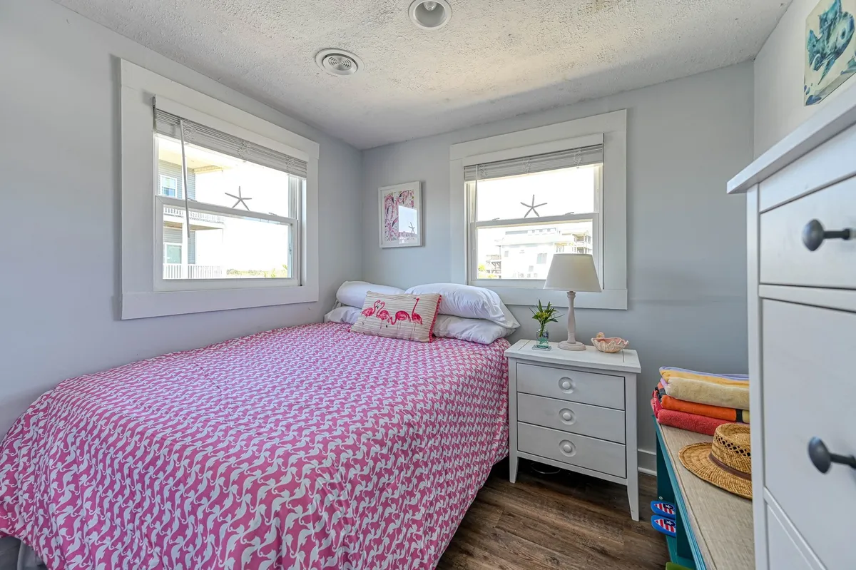 A bright and inviting bedroom featuring a bed dressed in cheerful pink patterned bedding, a white bedside table topped with a lamp, and decorative wall art. Sunlight pours in, creating a cozy and restful atmosphere that nods to vintage charm.