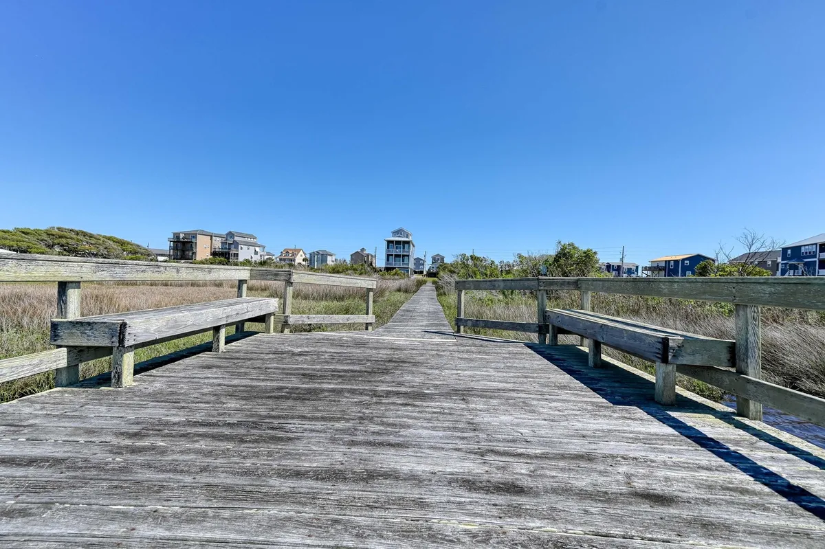 A wooden walkway stretches across marshland, with benches offering a place to relax and take in the scenery. Houses in the distance hint at a community vibe while the dock provides an excellent vantage for birdwatching and sunset views.