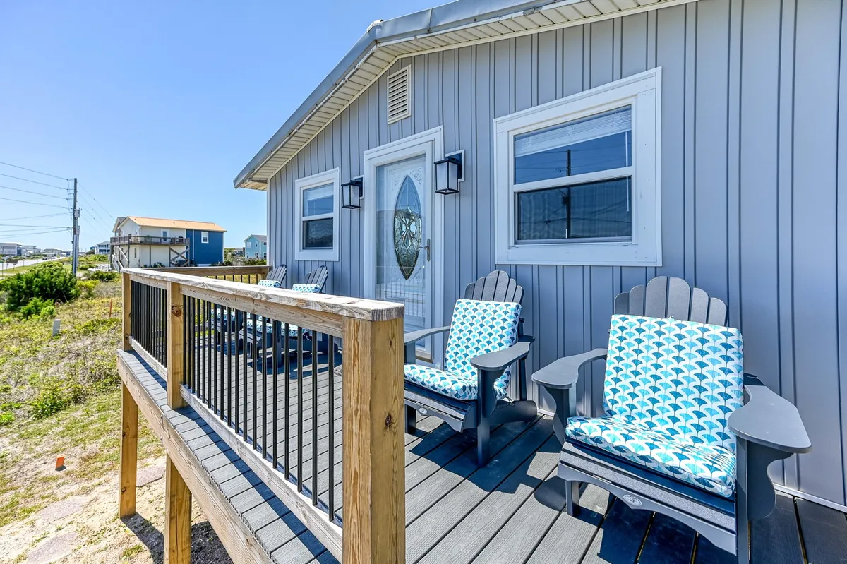 The image features blue and white patterned chairs arranged on a welcoming porch, set against a front door with decorative glass and a wooden railing. The space overlooks the coastal landscape, creating a serene spot perfect for morning coffee or sunset relaxation.