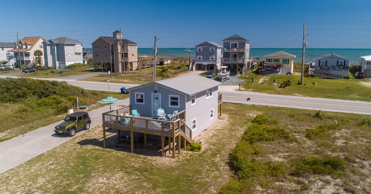 The hero image displays a charming vintage beach cottage with a welcoming deck furnished with chairs, facing the serene North Topsail shoreline. The beach is just steps away, surrounded by a peaceful neighborhood of coastal homes, all set beneath open skies that hint at beautiful sunrises and relaxing evenings.