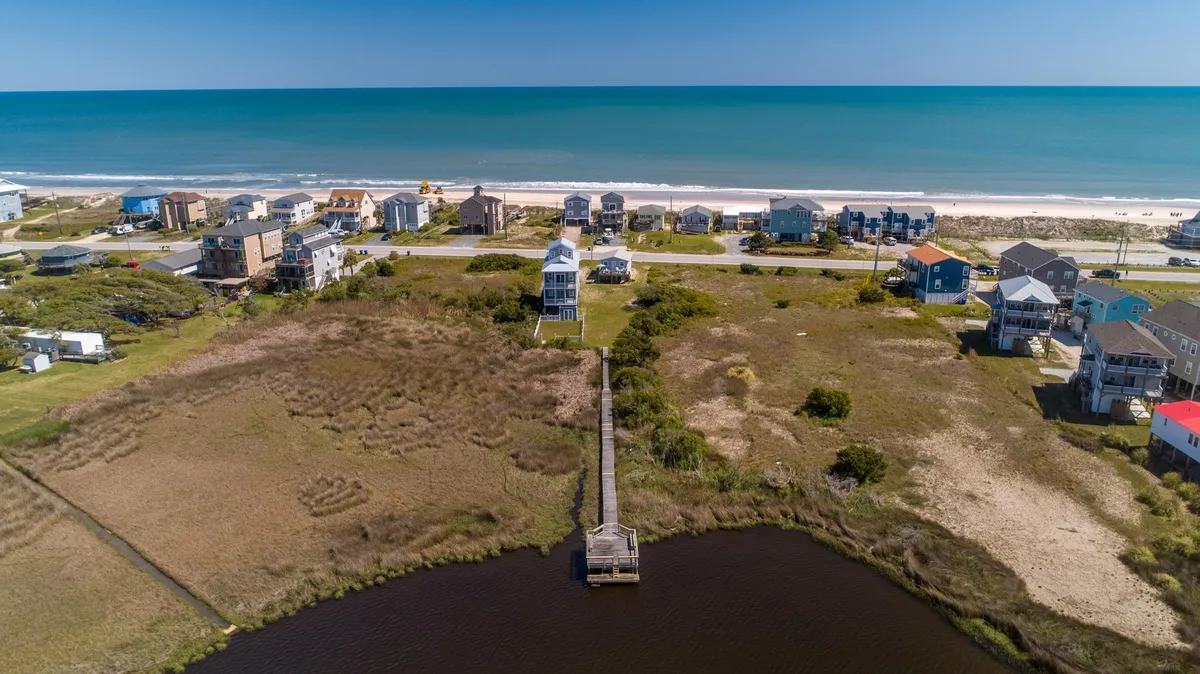 A sweeping view of the beachfront with a pier extending into the ocean, surrounded by coastal houses. The sandy beach is prominent, with the vast ocean stretching out towards the horizon, inviting beach activities and water adventures.