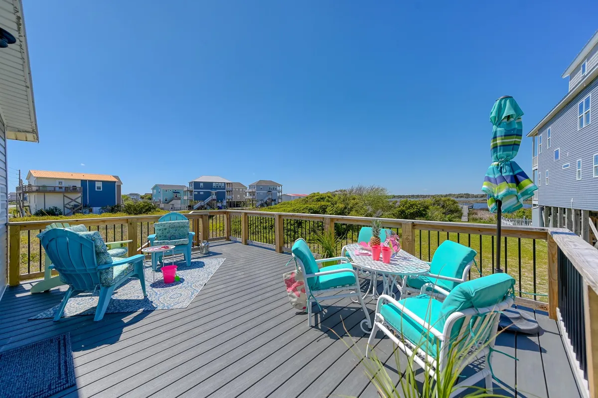 A vibrant and cheerful deck scene, this image features colorful outdoor furniture, a decorative table set with drinks, and a large parasol. The space is set for outdoor dining or casual lounging, with sunlight streaming in, creating an atmosphere ideal for al fresco meals and sunset relaxation.