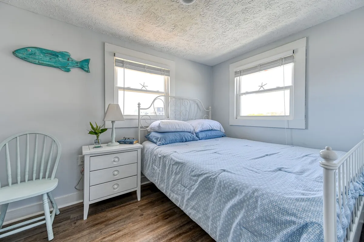 A serene bedroom with a bed featuring soft blue bedding, a white nightstand and lamp, decorative fish accents on the wall, and a window filling the space with natural light. The setting is calm and inviting, perfect for unwinding after a long day.